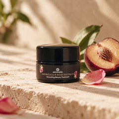 A small black jar of Annmarie Boswellia & Rose Eye Balm, with a plum and rose petal on a textured beige surface.