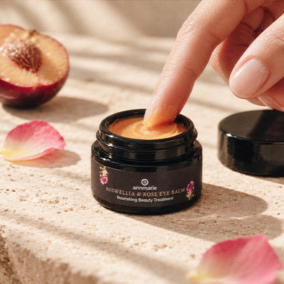 A close-up of a hand dipping a finger into a small black jar of Annmarie Boswellia & Rose Eye Balm The jar sits on a textured beige stone surface with scattered pink rose petals, a halved peach, and the black lid resting beside it.