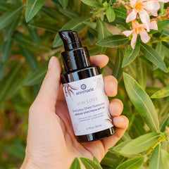 Hand holding Sun Love Everyday Sheer Sunscreen SPF 20 bottle surrounded by green leaves and soft pink flowers.